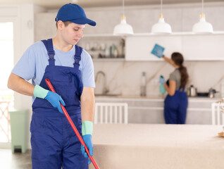 Focused skilled young guy from cleaning company wearing blue overalls, meticulously washing and mopping indoor floors