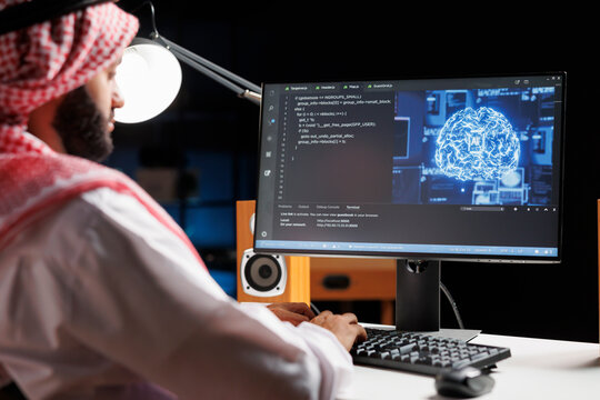 Muslim engineer focuses on a computer screen displaying code and data in a futuristic work setting. He manages a robust cloud-based system while monitoring data on a desktop computer screen.