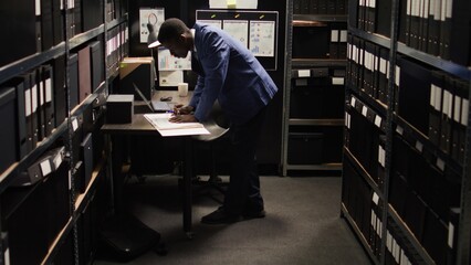 African American policeman carefully analyzes material, does records searches, and collects data from shelves and laptop. Male police officer standing, looking through case files on desk in office. - obrazy, fototapety, plakaty