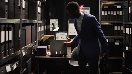 Male private investigator exits office room after packing his personal computer and closing it. African american police officer setting up his desk, organising paperwork, and carrying his laptop bag. - obrazy, fototapety, plakaty