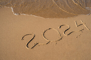 New Year concept. Text 2024 handwritten in sand surface. Blue ocean wave washing away numbers at the beach. Welcoming year 2024 Happy New Year coming new resolutions, dreams and goals