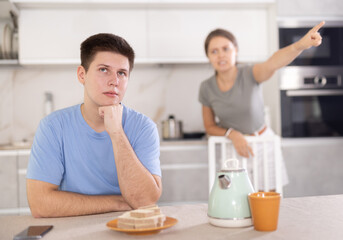 Frustrated offended guy sitting at kitchen table on background of swearing dissatisfied young wife. Family conflicts and communication problems concept