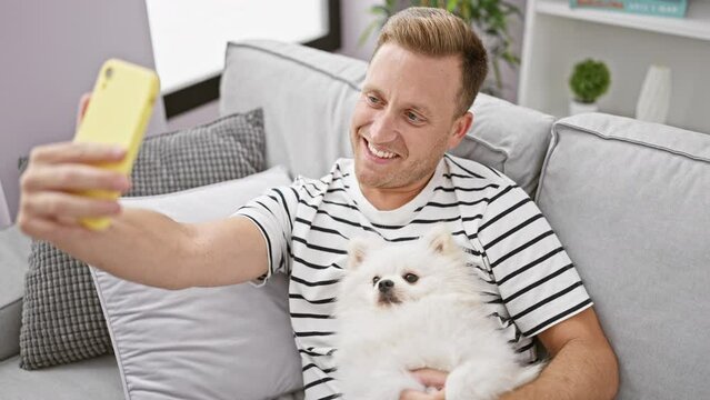 Young caucasian man with dog taking selfie picture with smartphone sitting on the sofa at home