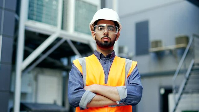 Portrait of confident professional engineer wearing safety helmet, protective glasses and vest standing at factory with crossed arms. Headshot of serious industry manager in uniform looking at camera