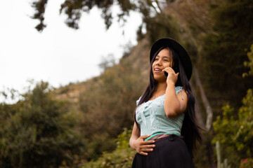 Foto de una feliz y bonita campesina caucásica con un sombrero hablando por un teléfono celular en los Andes peruanos. Mujer usando teléfono móvil - obrazy, fototapety, plakaty