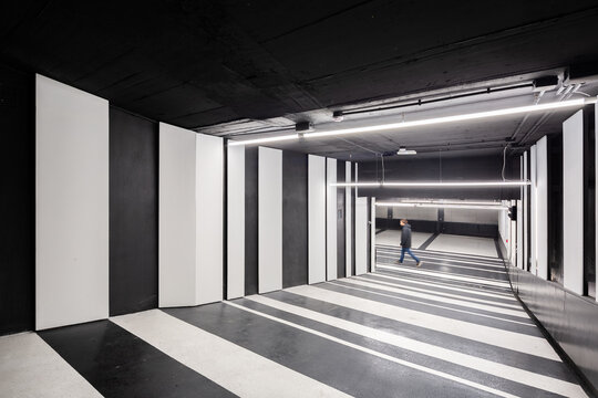 Pedestrian walking through a modern parking garage with striking black and white striped walls and floor patterns, illuminated by overhead fluorescent lights