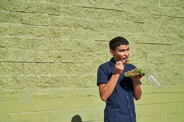 Young man eating Thai salad by green cinder block wall outdoors
