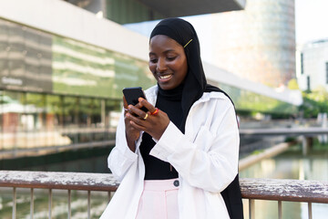 Cheerful African American young businesswoman in hijab browsing on smart phone while standing against modern building