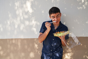 Young man eating Thai salad by green cinder block wall outdoors