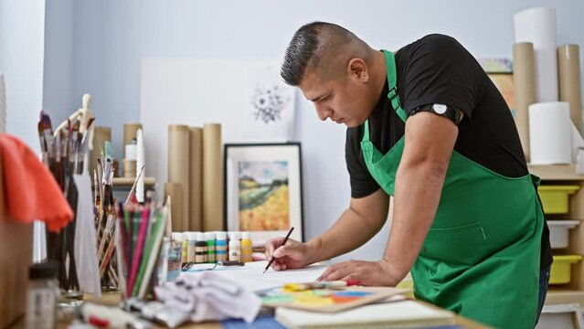 Handsome young latin artist engrossed in his drawing lesson indoors at art studio, standing at desk with canvas and paintbrushes for his passionate hobby