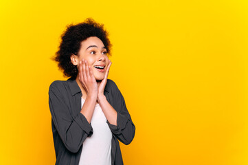 Excited surprised curly haired african american stylish positive woman, looks in amazement to the side at empty space, stands on isolated yellow background, smiling