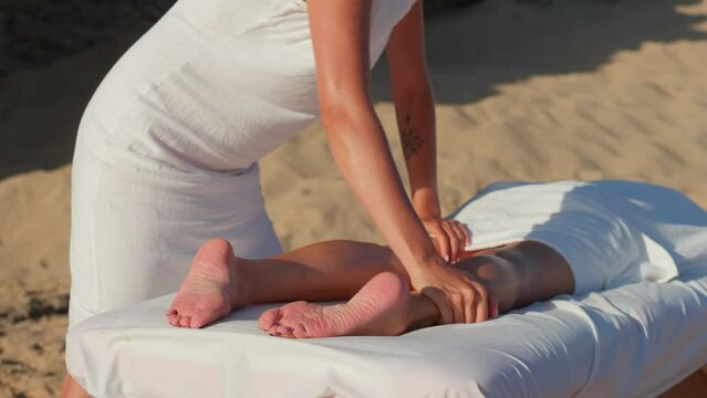 Female masseuse giving lymphatic drainage massage of legs and lower legs. Professional masseur giving spa massage client lying on special table standing on sandy beach. Healthy lifestyle and body care
