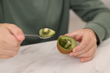 Boy eating tasty fresh kiwi with spoon at white marble table indoors, closeup