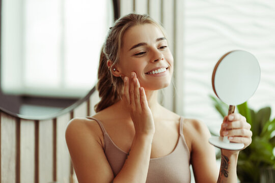 Attractive confident woman holding and looking in mirror wearing tank top standing in bathroom