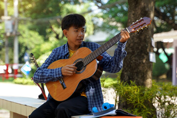 Asian boy happily sits outside playing guitar with his camp friends during break. Soft and...