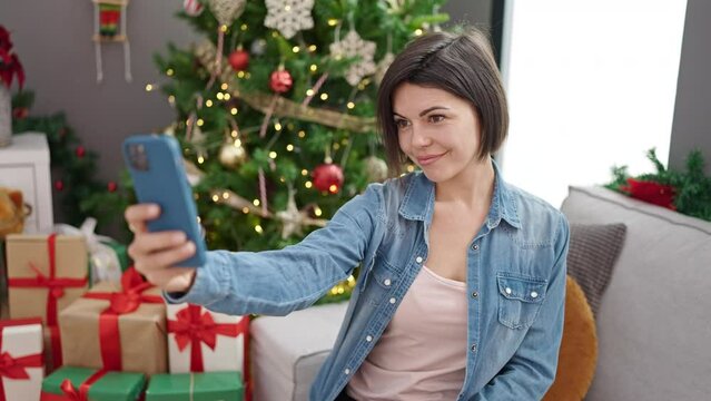 Young caucasian woman taking selfie with smartphone standing by christmas tree at home