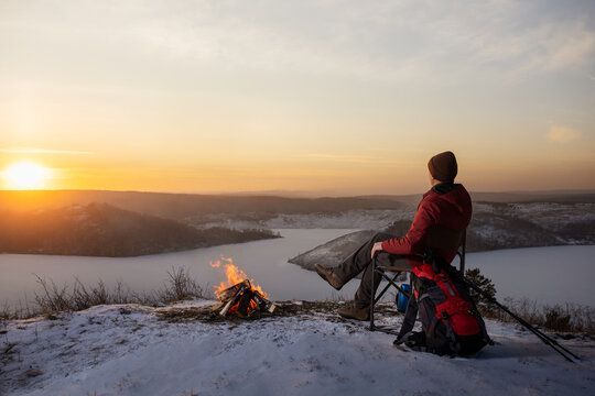  A man hiker sitting on the top of the mounting near campfire and enjoying yellow sunset at winter nature.  Travel, lifestyle, freedom concept.