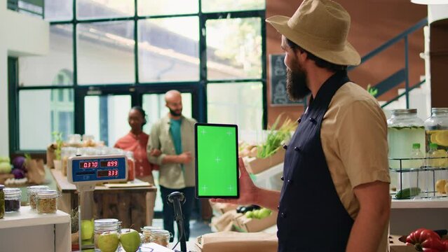 Farmer holding tablet with greenscreen in zero waste eco market, using copyspace template on mobile device standing at cash register counter. Small business owner presents isolated display.