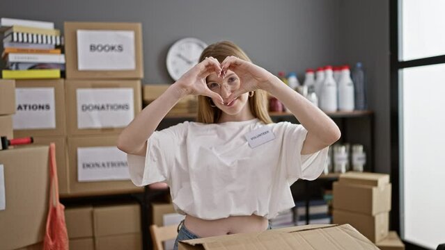 Young blonde woman volunteer smiling confident doing heart gesture at charity center