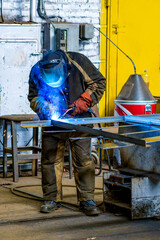 A welder wearing a protective mask for metal welding and protective gloves performs welding work at a metal structures plant. Welding a steel industrial beam. Sparks, hot flame from welding.