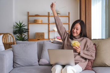 Portrait image of a happy woman eating potato chips while working or watching movie on laptop at home - Powered by Adobe