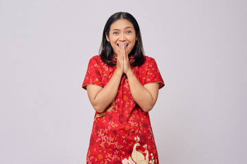 Excited young Asian woman wearing a traditional cheongsam qipao dress covering mouth with hand and looking at camera isolated on white background. Happy Chinese New Year