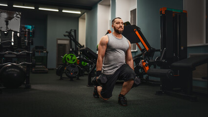 A man with a beard athlete performs an exercise lunges with dumbbells in a large gym for...
