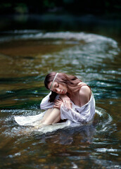 Fototapeta premium Young long haired woman in slavic style white nightgown with open shoulders and hair band sitting in river in dark forest. Tradinional Ivan Kupala outfit. Fairytale portrait of a girl