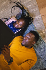 Happy african american father and daughter lying on floor and using tablet at home, copy space