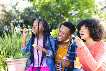 Happy african american father, mother and daughter blowing bubbles in sunny garden, copy space