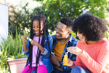Happy african american father, mother and daughter blowing bubbles in sunny garden, copy space