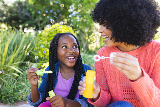 Happy african american mother and daughter sitting and blowing bubbles in sunny garden, copy space