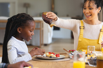 Happy african american mother pouring maple syrup on fruits in dining room at home, copy space - obrazy, fototapety, plakaty