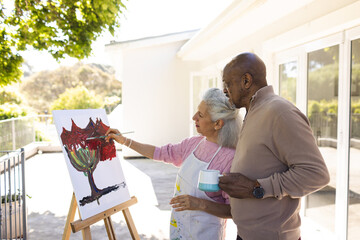 Diverse senior woman painting on canvas and senior man holding cup of coffee on sunny terrace