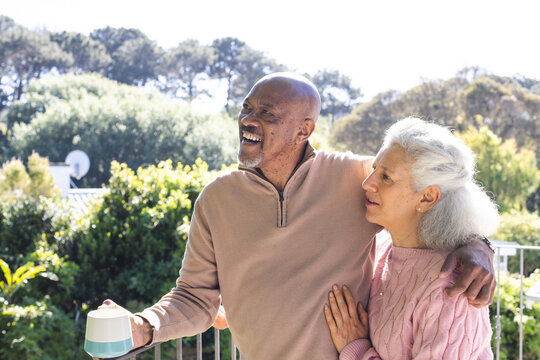 Happy diverse senior couple holding cup of coffee and embracing on sunny terrace