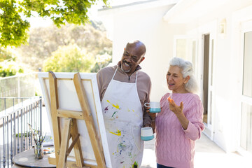 Happy diverse senior couple drinking coffee, embracing and looking at painting on sunny terrace