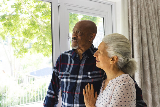 Diverse senior couple embracing and looking out window at sunny home