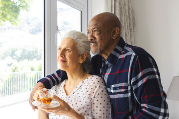 Diverse senior couple drinking tea, embracing and looking out window at sunny home