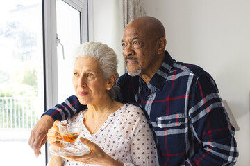 Diverse senior couple drinking tea, embracing and looking out window at sunny home