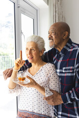 Diverse senior couple drinking tea, embracing and looking out window at sunny home