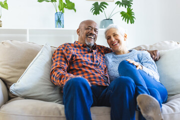 Happy diverse senior couple sitting on sofa and embracing in sunny living room - Powered by Adobe