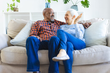 Happy diverse senior couple sitting on sofa, talking and embracing in sunny living room