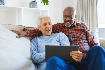 Diverse senior couple sitting on sofa, talking, embracing and using tablet in sunny living room - Powered by Adobe