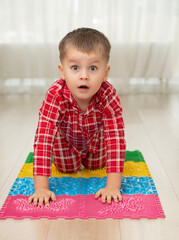 Sport and health concept. A little boy 4 years old in red checkered pajamas is working out on a multi-colored massage orthopedic mat with spikes in a home interior. Close-up.