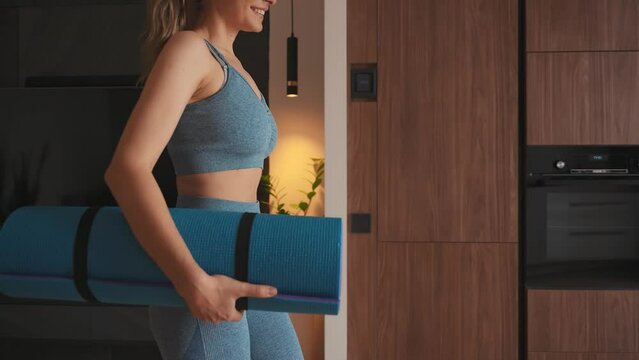 young happy woman going to do sports at home back rear view, holds karimat mat in hands, sportswear suit blue top leggings. modern room interior. aerobics sport fit yoga workout domestic fitness 4k