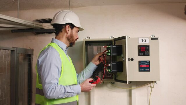 Serious electrician with electrical tester inspecting switchboard functionality at power substation, technician in protective outfit examining voltage on transmission lines in factory technical area.