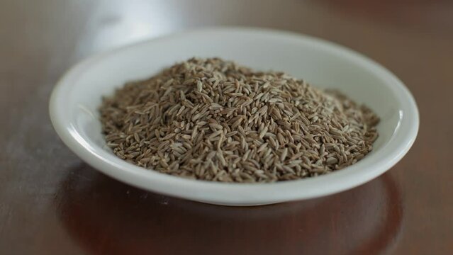 Brown crumbly spice cumin lies in a white deep plate. The product for preparing lunch, cumin, is at hand for the cook. Smooth departure and arrival at delicious aromatic cumin.