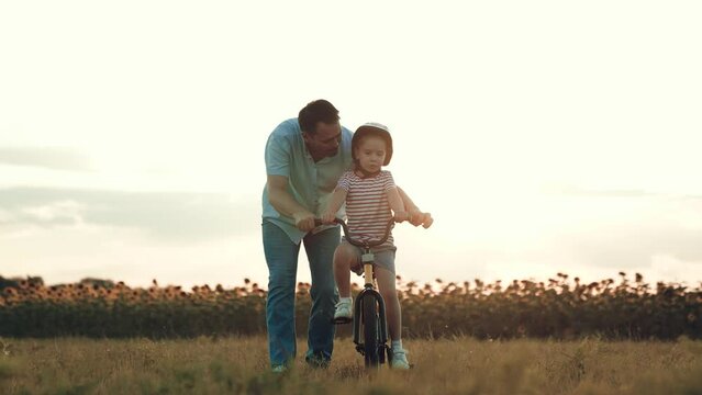 Father holds bicycle while little daughter learns ride vehicle in rural meadow