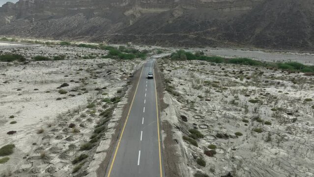 Aerial drone backward moving shot over a car moving along a highway in Hingol National Park with mountain range in the background in Balochistan on a cloudy day.