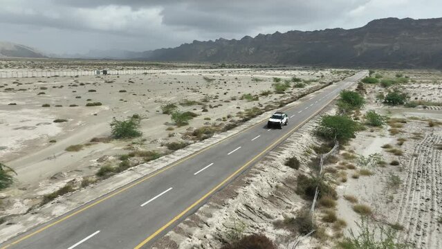 Aerial drone following shot over a car driving along the highway road through Hingol National Park in Balochistan on a cloudy day. Desert vegetation.